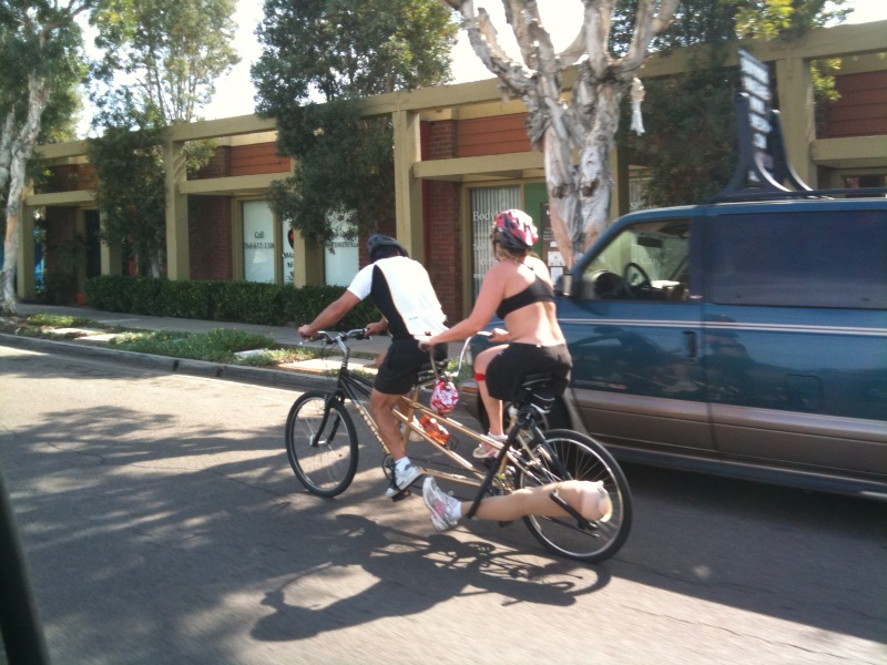 two people on a tandem bike with only 3 legs; two in front, one in back